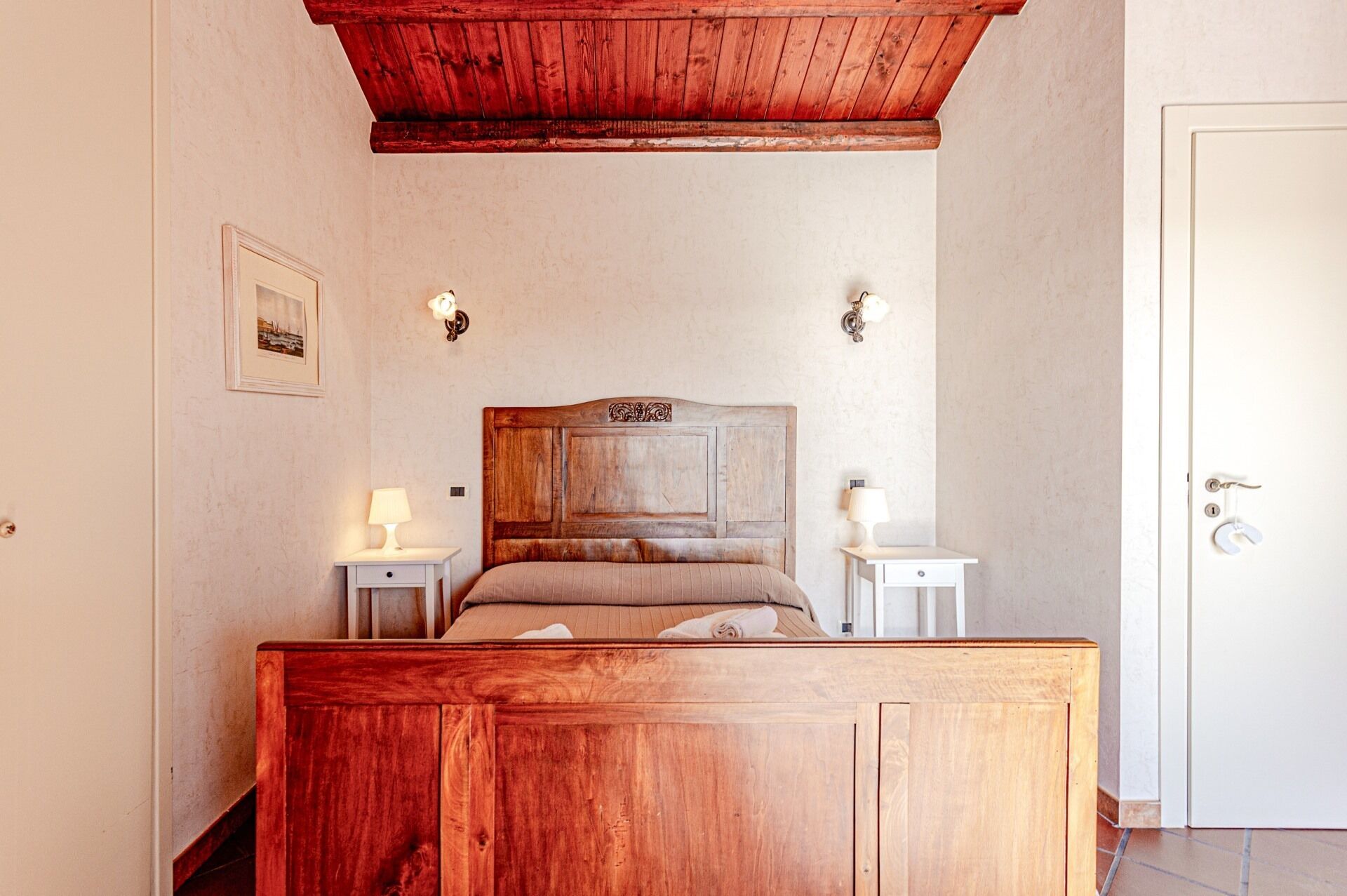 A bedroom with a wooden headboard, two bedside tables with lamps, a framed picture on the wall, and a door with a key in the lock.