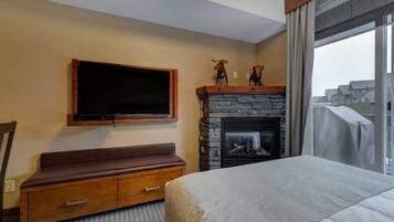 A hotel room with a stone fireplace, a large flat-screen TV mounted on the wall, a wooden bench, and a bed.