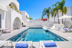 A swimming pool with lounge chairs and umbrellas, surrounded by white buildings and palm trees.