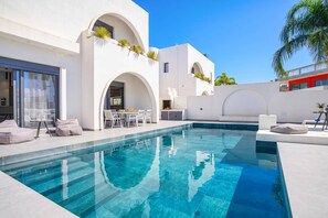 Un área de piscina moderna con piscina azul cristalina, paredes blancas y un comedor con asientos al aire libre.