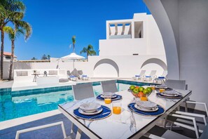 A poolside dining area with a table set for two, a pool, and a white building with arched windows in the background.