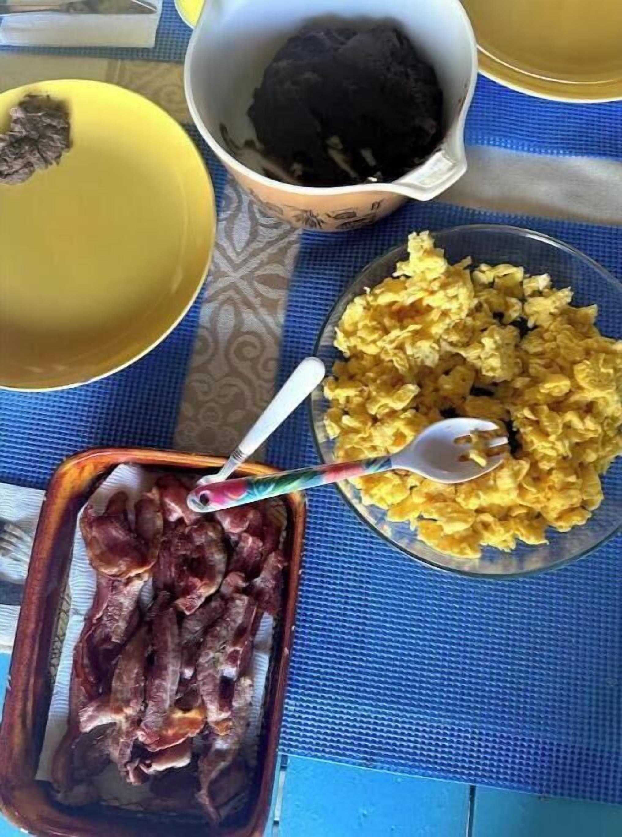 A table with a variety of breakfast items including bacon, scrambled eggs, and a bowl of black pudding.