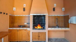 Een keuken met een centraal geplaatste open haard, een betegelde spatwand, houten kastjes en een spoelbak.