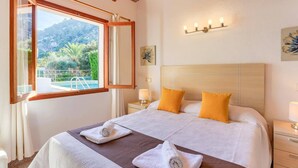 A bedroom with a large bed, two pillows, a nightstand with a lamp, and a window with a view of a pool and greenery. - Villa Rabasso by Villa Plus (Pollença)