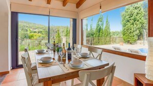 A dining area with a table set for four, large windows offering a view of a green landscape and a pool. - Villa Marinada by Villa Plus (Pollença)