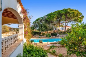 A pool area with a clear view of a building with an arched entrance, surrounded by lush greenery and trees. - Villa Cas Sucrer by Villa Plus (Binibeca)