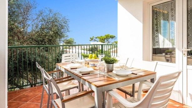 A balcony with a table set for two, surrounded by white chairs, a green railing, and a view of trees and buildings.