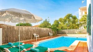 A poolside area with lounge chairs and a covered umbrella, a stone wall, and a building in the background.