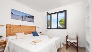 Une chambre avec une tête de lit en bois, deux lits avec du linge de lit blanc, une grande fenêtre donnant sur la mer et une chaise en bois.