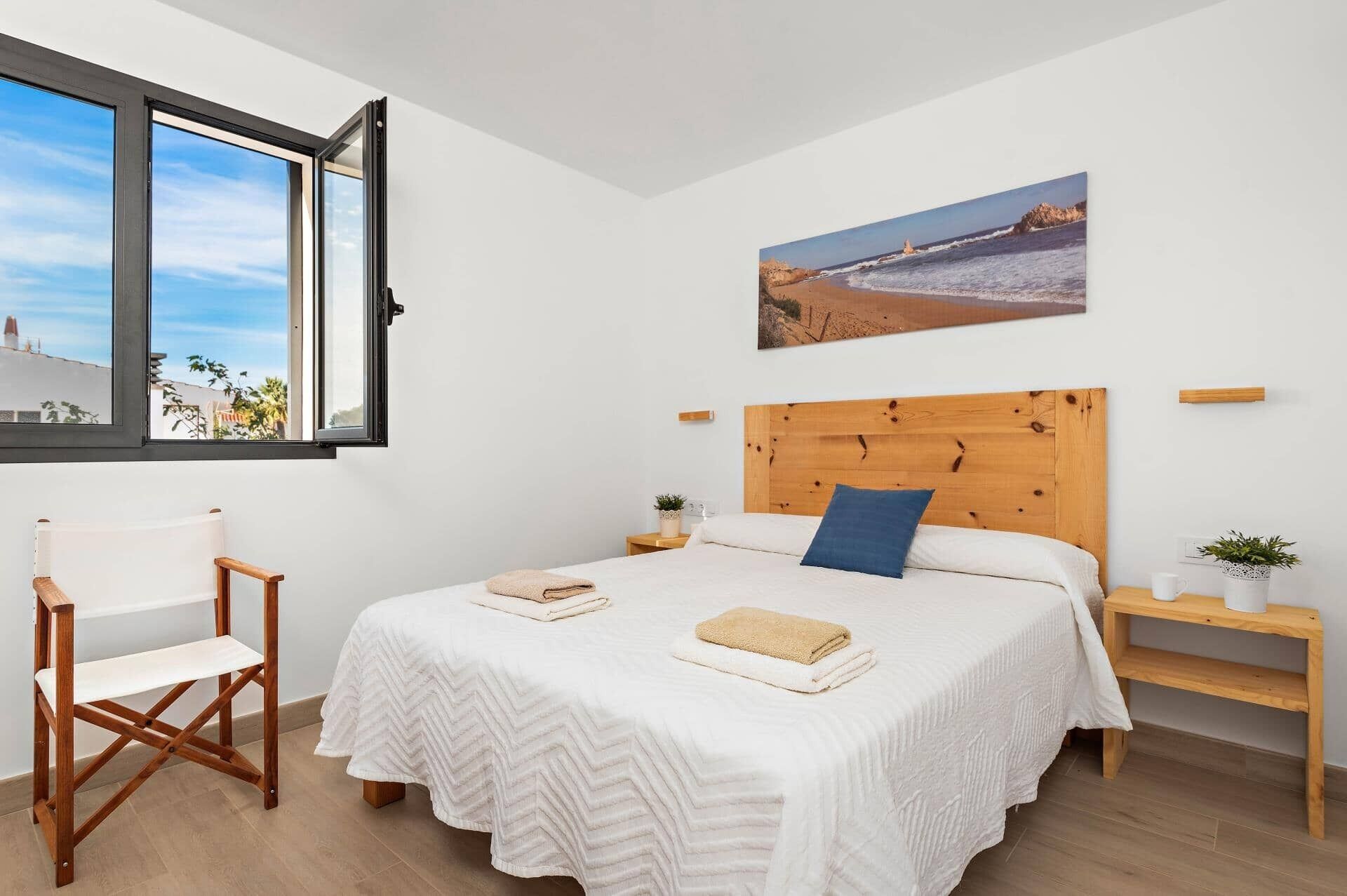 A bedroom with a large bed, a wooden headboard, a window with a view of the sea, a painting of a beach, and a wooden chair.