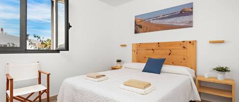 A bedroom with a large bed, a wooden headboard, a window with a view of the sea, a painting of a beach, and a wooden chair.