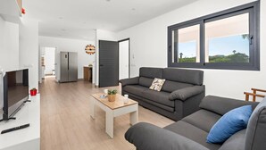 Un salon moderne avec un canapé gris, une table basse et un téléviseur. On aperçoit une cuisine au fond, avec un réfrigérateur et une horloge au mur.