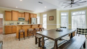 A kitchen with wooden cabinets, a central island with stools, a ceiling fan, and a large window with blinds. - Lake Hartwell Getaway w/ Private Dock + Fire Pit! (Fair Play)