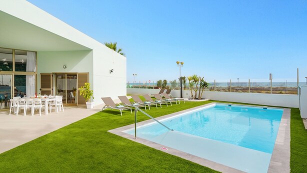 A modern outdoor pool area with a clear view of the ocean, a dining area with white chairs, and a well-maintained lawn.