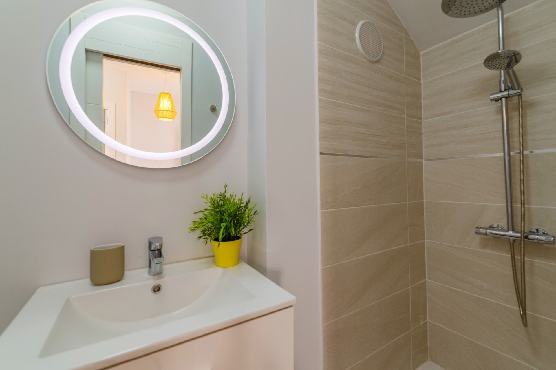 Une salle de bain avec un miroir rond, un lavabo, une plante en pot et une douche équipée d’un pommeau de douche à main.