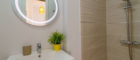 Une salle de bain avec un miroir rond, un lavabo, une plante en pot et une douche équipée d’un pommeau de douche à main.