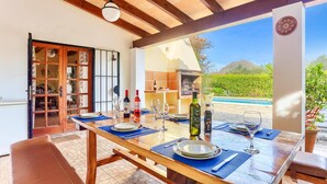Un área de comedor al aire libre con una mesa de madera preparada para cuatro personas, rodeada de vistas a una piscina y un jardín.