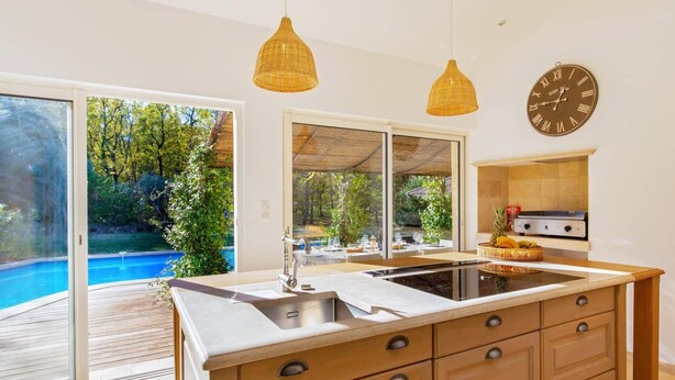 A modern kitchen with a large island, a built-in grill, and a view of a swimming pool. - Villa Beauregard by Villa Plus (Tourrettes)