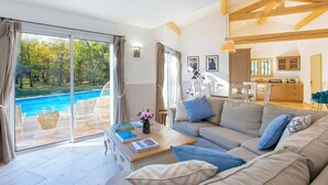 A spacious living room with a large sofa, a coffee table, and a view of a swimming pool. - Villa Beauregard by Villa Plus (Tourrettes)
