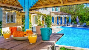 A poolside breakfast setup with a table, orange juice, croissants, and a turquoise pitcher. - Villa Beauregard by Villa Plus (Tourrettes)