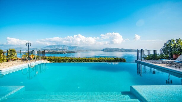 A clear blue infinity pool with a view of a lake and distant mountains, surrounded by a metal fence and potted plants.