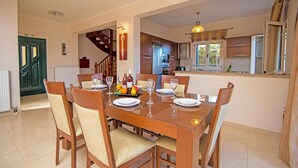 A dining area with a wooden table set for six, surrounded by chairs. A kitchen is visible in the background with modern appliances.