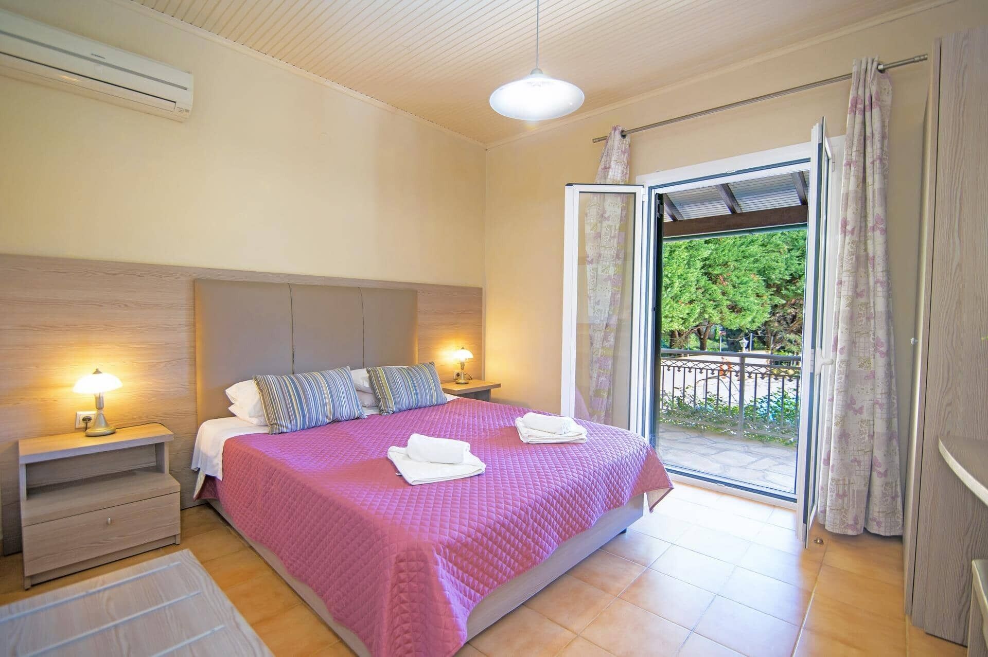 A bedroom with a bed, nightstands, a lamp, and a view of the outdoors.
