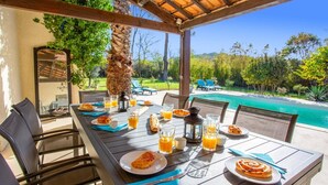 Tavolo apparecchiato all'aperto con cibi per la colazione e bicchieri di succo d'arancia, con vista su una piscina e vegetazione lussureggiante.