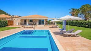 Une piscine avec des chaises longues et des parasols, une maison en pierre avec un toit en tuiles et un ciel d’un bleu limpide.