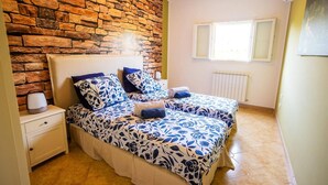 A bedroom with a stone wall, two beds with blue floral bedding, a bedside table with a lamp, and a window with a view.