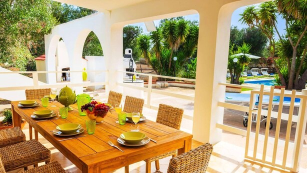 An outdoor dining area with a wooden table set for six, surrounded by wicker chairs, overlooking a pool and lush greenery.