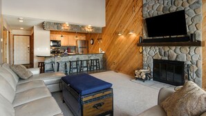 Ein gemütliches Wohnzimmer mit Kamin aus Stein, Holzboden und einer Küche im Hintergrund.