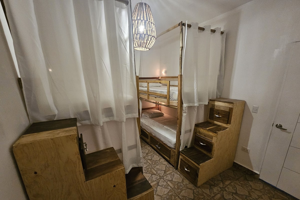 A dormitory room with bunk beds, a wooden wardrobe, a small desk, and a hanging light fixture.
