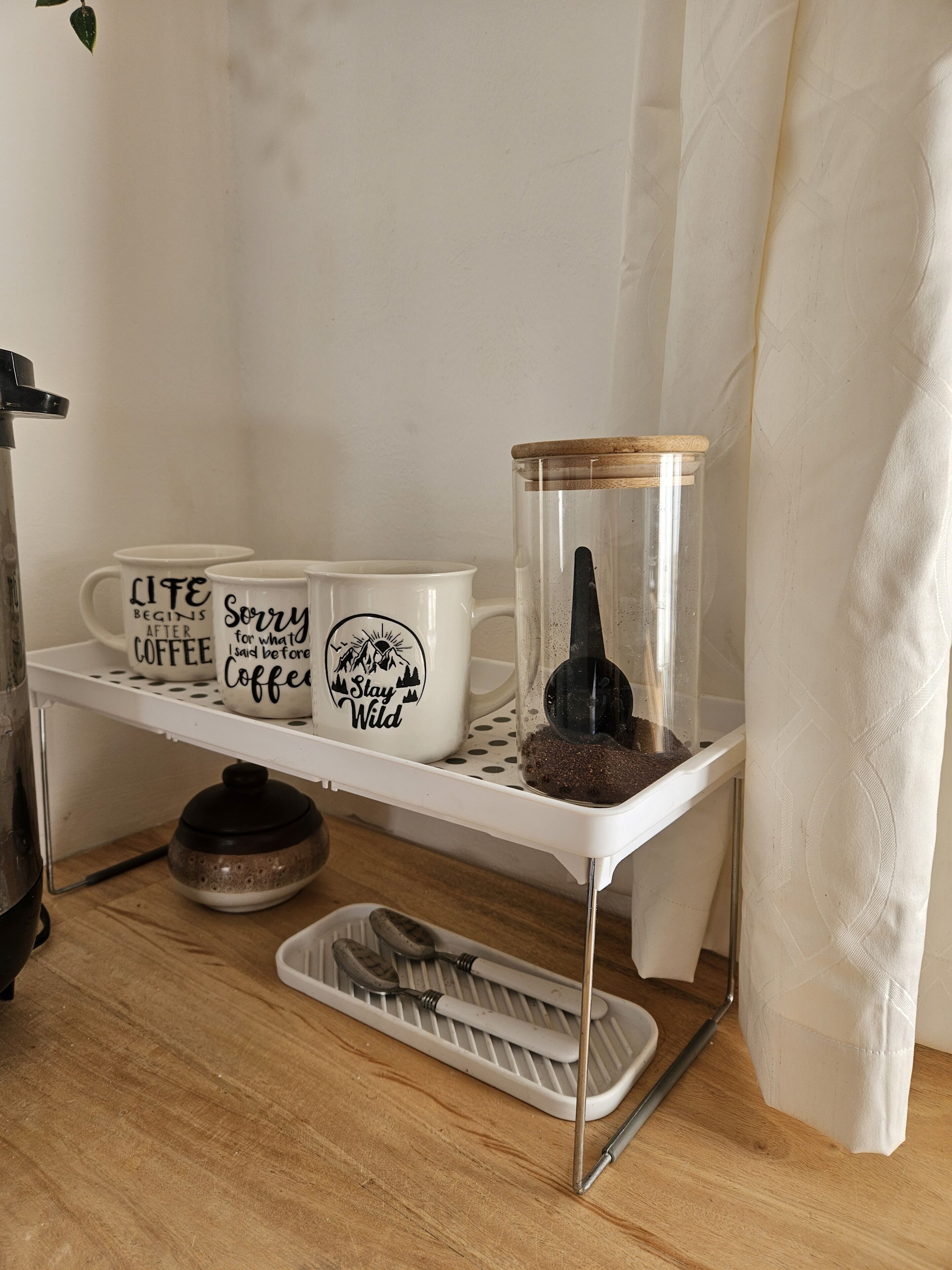 A kitchen counter with a coffee maker, two mugs with coffee-related phrases, a glass jar with a plant, and a tray with utensils.