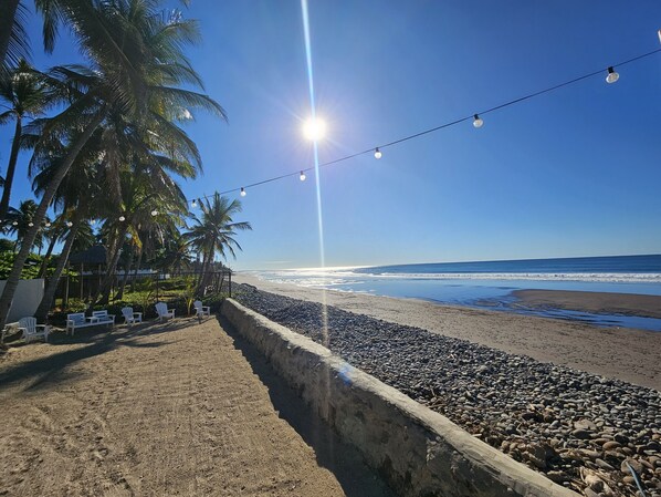 A beach with a concrete wall, white chairs, and palm trees under string lights. - Sunset Surf Villa (La Libertad)