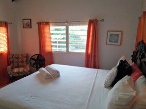 Una cama bien hecha con una toalla doblada, una silla con un cojín estampado, un ventilador de pie y una ventana con cortinas.