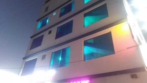 Un immeuble à plusieurs étages, avec des fenêtres illuminées et un panneau néon sur lequel on peut lire « S.A.V TOP ».