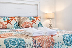 A neatly made bed with a floral and starfish patterned bedspread, a pair of folded white towels, and a bedside lamp on a white nightstand.