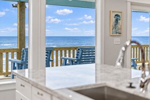 A kitchen with a view of the ocean, wooden deck chairs, and a painting on the wall.