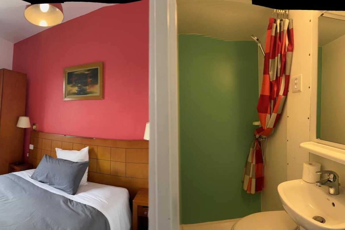 A hotel room with a bed, a painting on the wall, a window with curtains, a radiator, a wardrobe, and a bathroom with a sink and toilet.