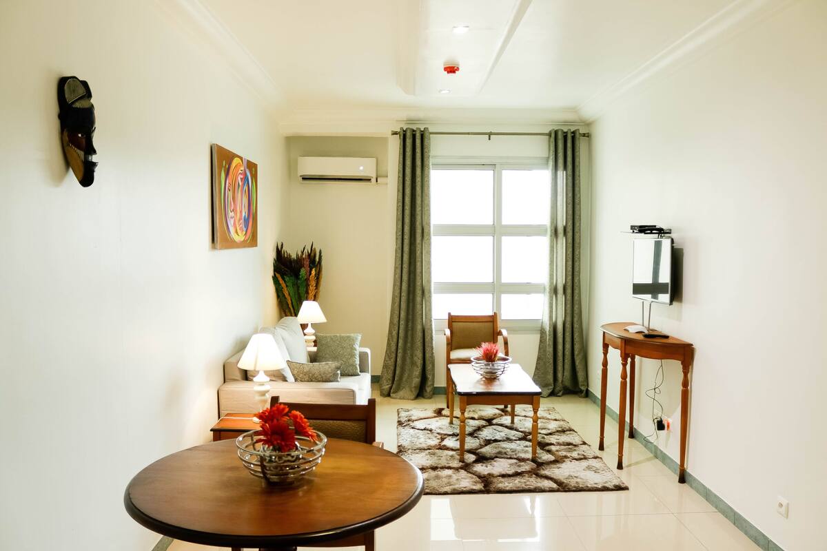Una sala de estar con un sofá, una mesa de centro con un florero, un televisor sobre un mueble bajo y una ventana con cortinas.