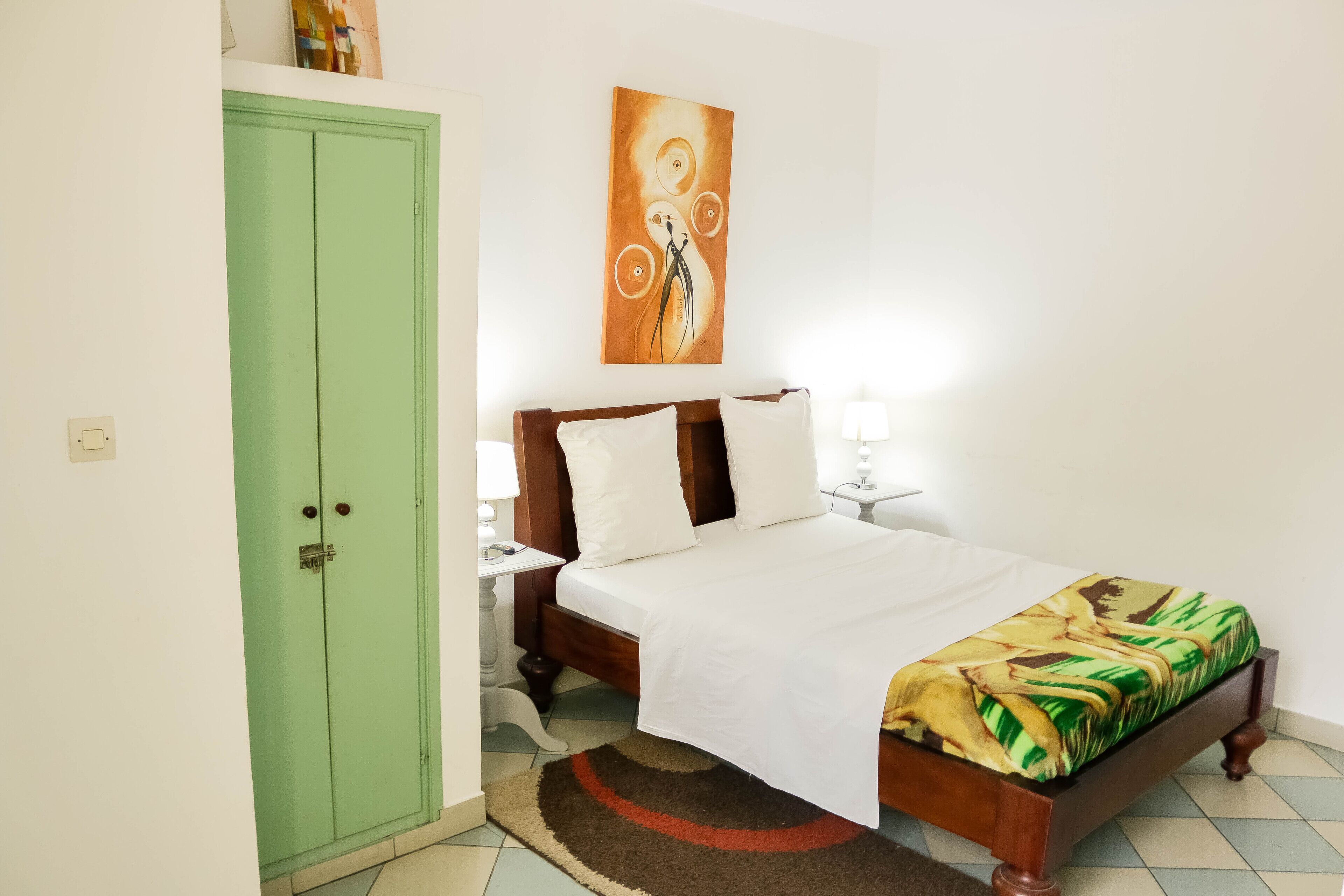 A bedroom with a green wardrobe, a bed with a white bedspread and a patterned blanket, a nightstand with a lamp, and a wall-mounted painting.