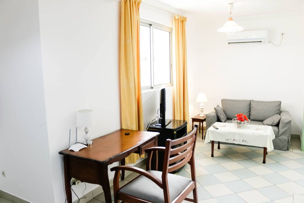 Una habitación pequeña y bien iluminada con un escritorio de madera, televisor, sofá y una mesa con un florero.