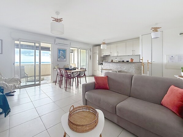 A modern living room with a grey sofa, a dining area with a table and chairs, and a kitchen with white cabinets.