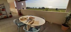 Et rundt bord med stole på en balkon, en potteplante og udsigt til havet.