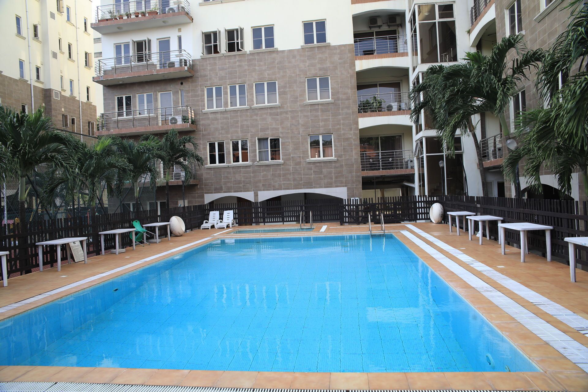 A swimming pool with a tiled deck, surrounded by a fence, and adjacent to a multi-story building with balconies.
