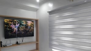 Una habitación moderna con un televisor montado en un estante de madera, una alfombra blanca y esponjosa y persianas horizontales en la ventana.