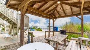 A wooden deck with a covered seating area, a white table, and a view of the beach and pier in the background. - Summer House West C202 (Gulf Shores)