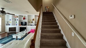 Una escalera conduce a una sala de estar con un sofá blanco, una mesa de centro y un televisor sobre un mueble blanco.