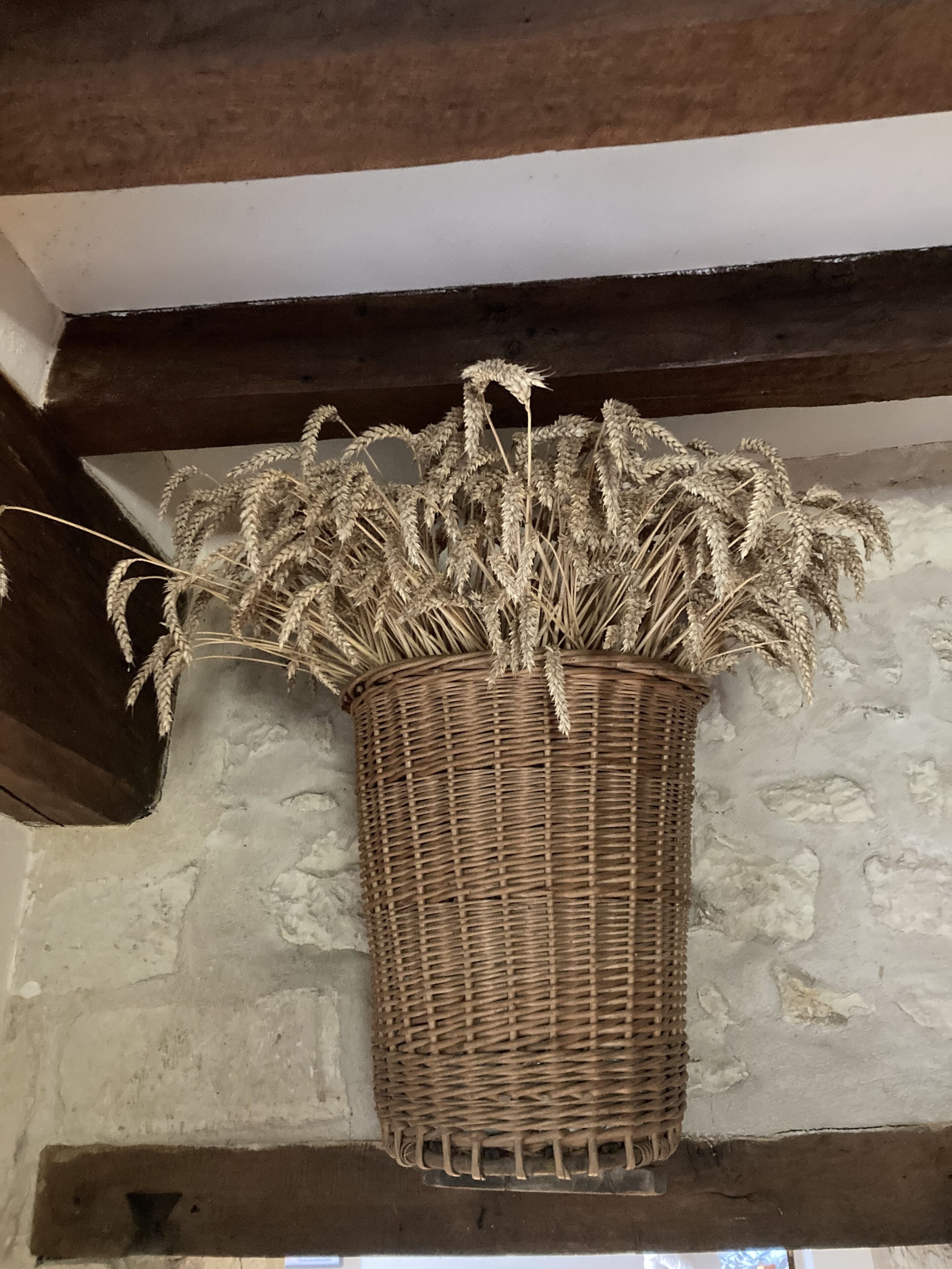 Una cesta de mimbre con espigas de trigo colgadas en una pared de piedra con vigas de madera.